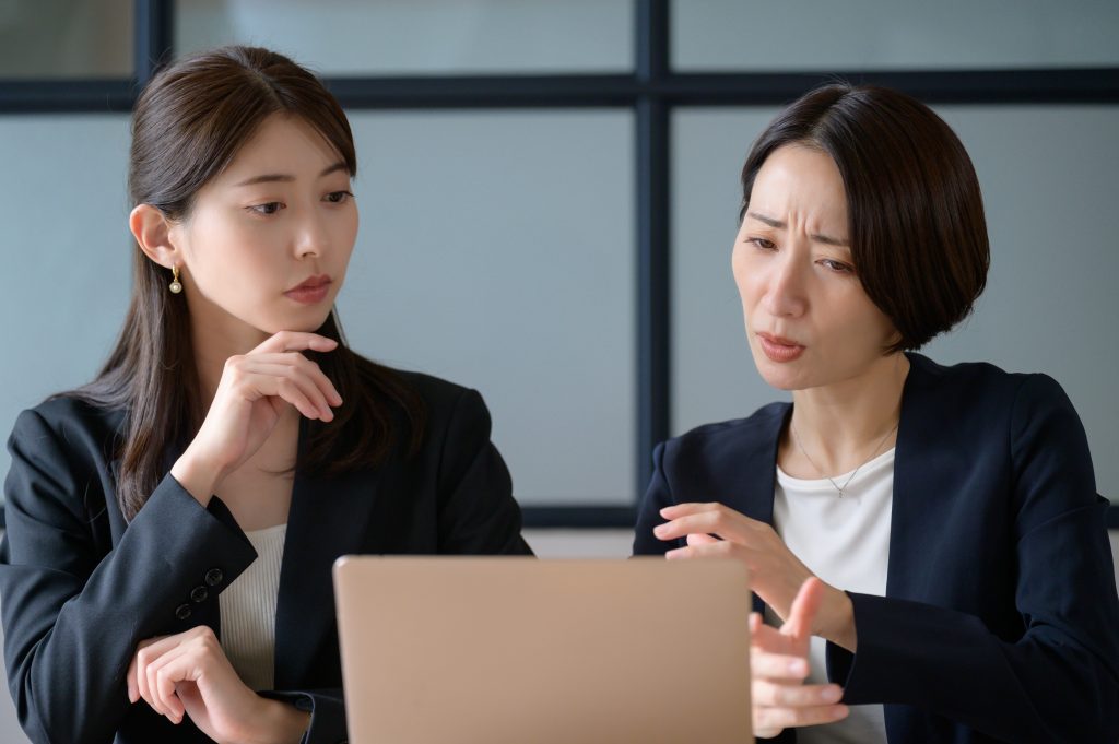 Excelやメールでのバラバラな来場者・講演者管理に限界を感じ、パソコンを前に困惑した表情で相談し合うイベント事務局のスタッフ
